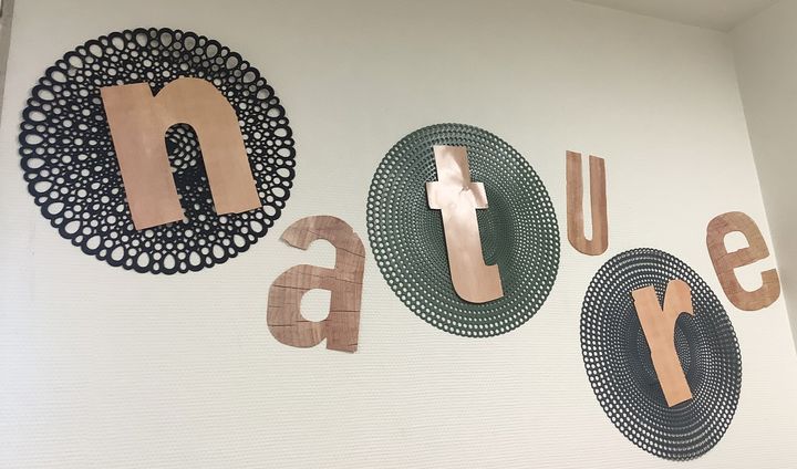 Le mot « nature » en lettres de bois et de cuivre, posé sur des sets ronds ajourés accrochés au mur.